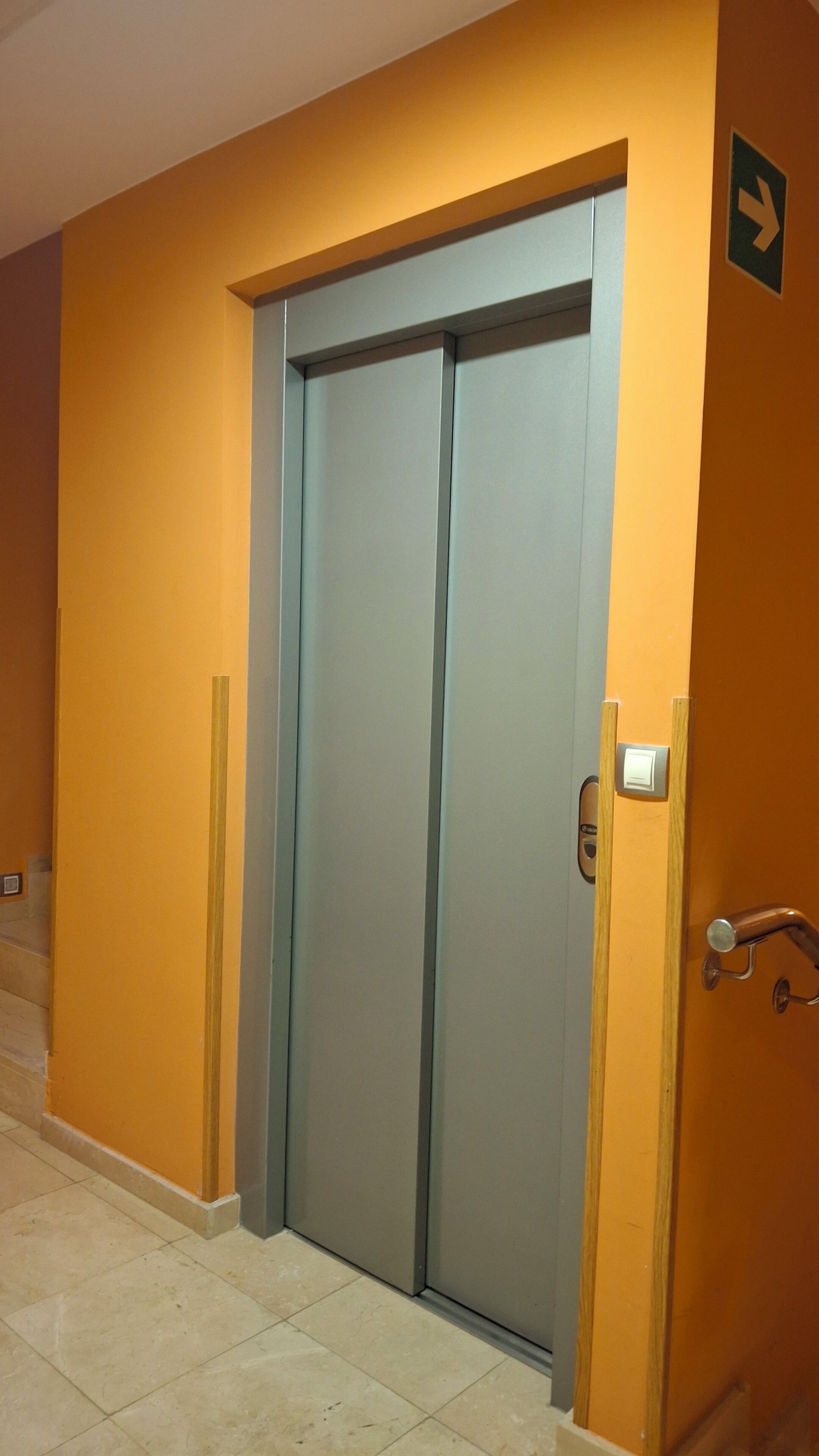 Puerta de ascensor cerrada con acabado metálico, un portero automático en la pared y un cartel indicador.