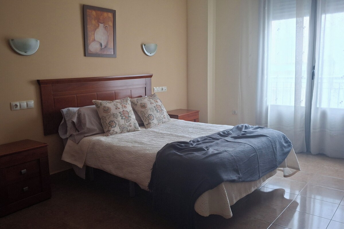 A bedroom with a bed, nightstands, a wooden headboard, a painting, and a window with curtains.