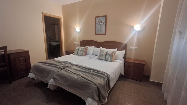 A hotel room with a large bed, two bedside tables, a nightstand, a door leading to a bathroom, and a wall-mounted light fixture. - Hostal Ruralis Altozano  (Montánchez)