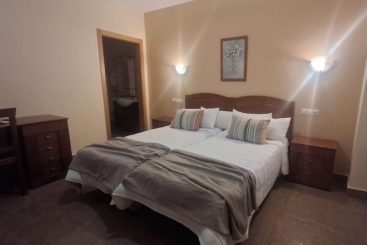 A hotel room with a large bed, two bedside tables, a nightstand, a door leading to a bathroom, and a wall-mounted light fixture.