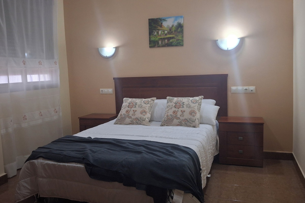 A bedroom with a bed, two bedside tables, a nightstand, a painting on the wall, and a window with curtains.