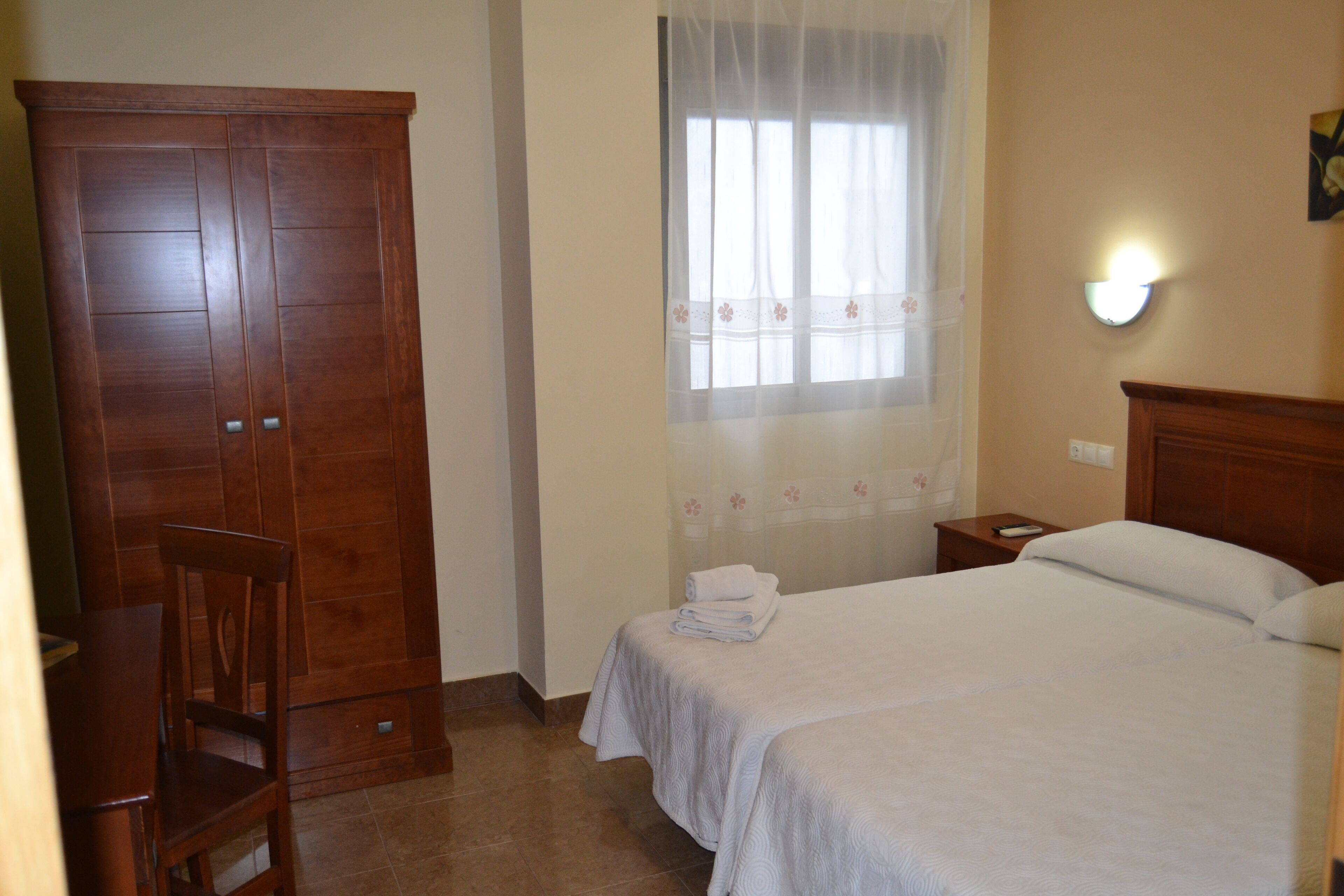 A bedroom with a bed, a wooden wardrobe, a chair, a nightstand, and a window with a sheer curtain.