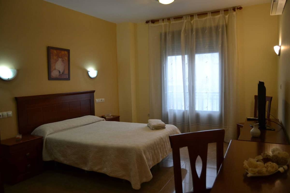 A hotel room with a bed, a chair, a table, a television, and a window with curtains.