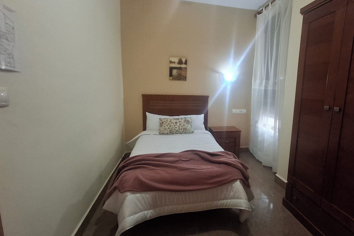 A hotel room with a bed, a nightstand, a wooden wardrobe, and a wall-mounted light fixture.