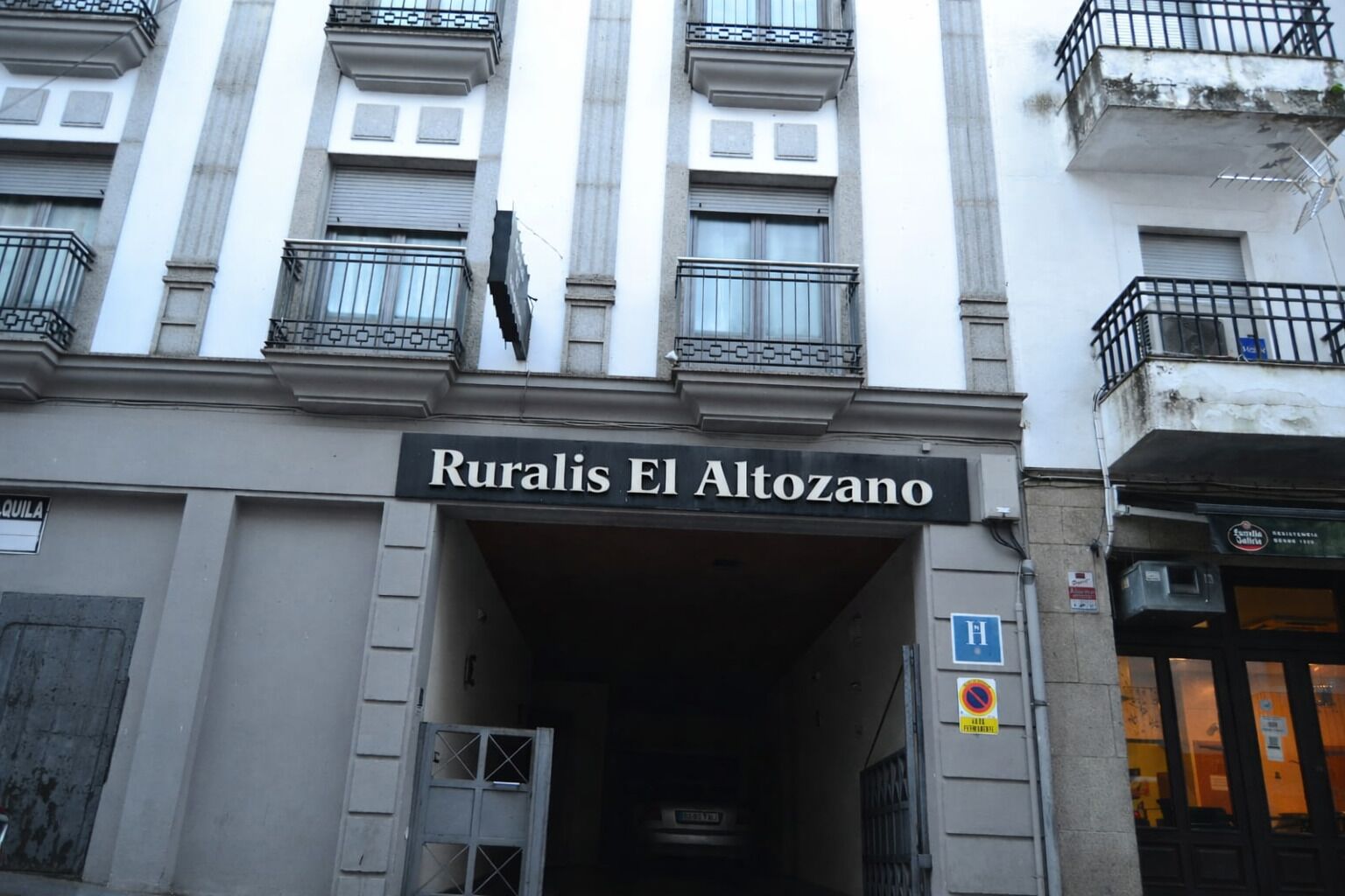 De ingang van Ruralis El Altozano, een gebouw met een zwart bord boven de ingang, een blauw parkeersbord en een verboden parkeren bord aan de rechterkant.