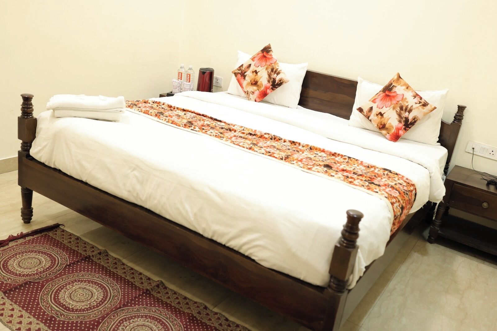 A neatly made bed with a floral-patterned bedspread and pillows, a wooden headboard, and a bedside table with a lamp and water bottles.