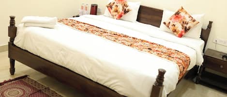 A neatly made bed with a floral-patterned bedspread and pillows, a wooden headboard, and a bedside table with a lamp and water bottles.