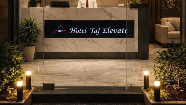 A reception desk at Hotel Taj Elevate with a receptionist, a clock, and a sign indicating the reception area. - Hotel Taj Elevate (Agra)
