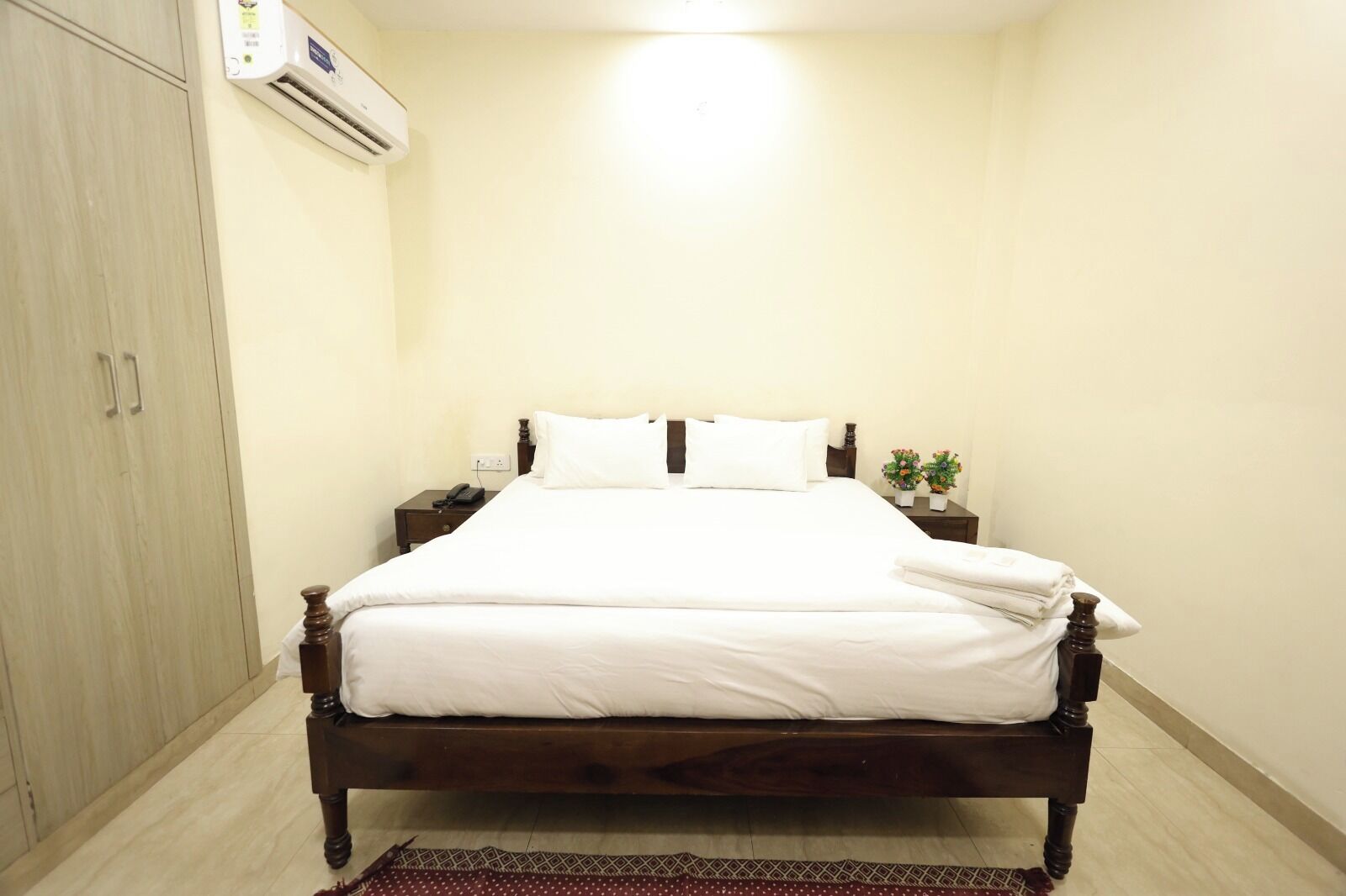 A neatly made bed with white linens, a wooden headboard, and a small bedside table with a phone and a plant.