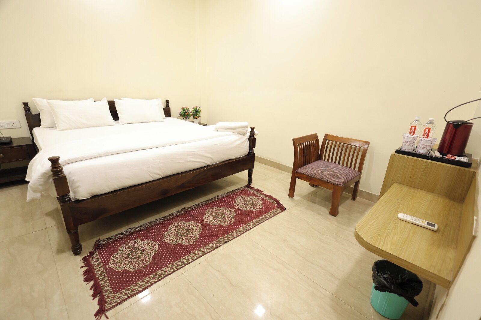 A neatly arranged bedroom with a wooden bed, a single chair, a small table with a remote control, and a trash bin.