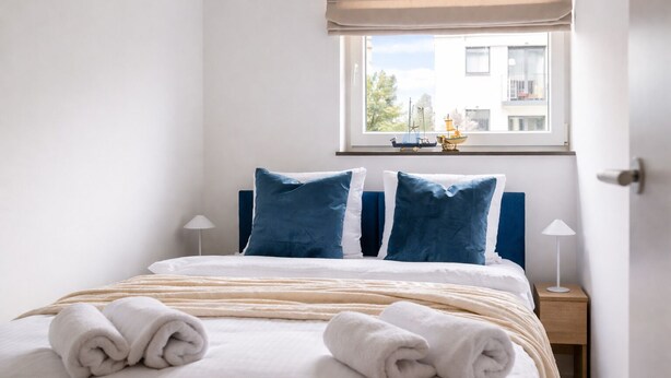 A neatly made bed with blue and white pillows, a window with a view of buildings, and a small table with a lamp. - HoliApart Trimare Billard &Seasonal Pool (Sztutowo)