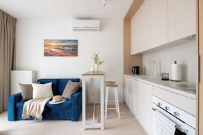 A modern kitchen with white cabinets, a white countertop, and a stove. There is a blue sofa with a beige blanket and a hat, and a small table with a plant. - HoliApart Trimare Billard &Seasonal Pool (Sztutowo)