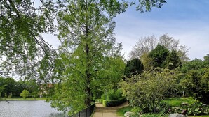 Un chemin pavé longe un plan d’eau, bordé d’une végétation luxuriante et d’arbres.