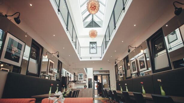 Un hall d’hôtel moderne avec un plafond haut, un espace salon central doté de canapés orange et un coin repas avec des chaises et des tables noires.