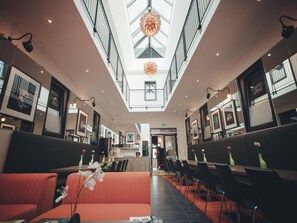 A modern hotel lobby with a high ceiling, a central seating area with orange couches, and a dining section with black chairs and tables.