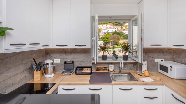 Una cucina moderna con mobili bianchi, una parete di fondo in marmo e vista su una zona residenziale attraverso una finestra aperta.