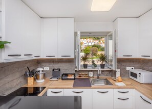 Una cucina moderna con mobili bianchi, una parete di fondo in marmo e vista su una zona residenziale attraverso una finestra aperta.