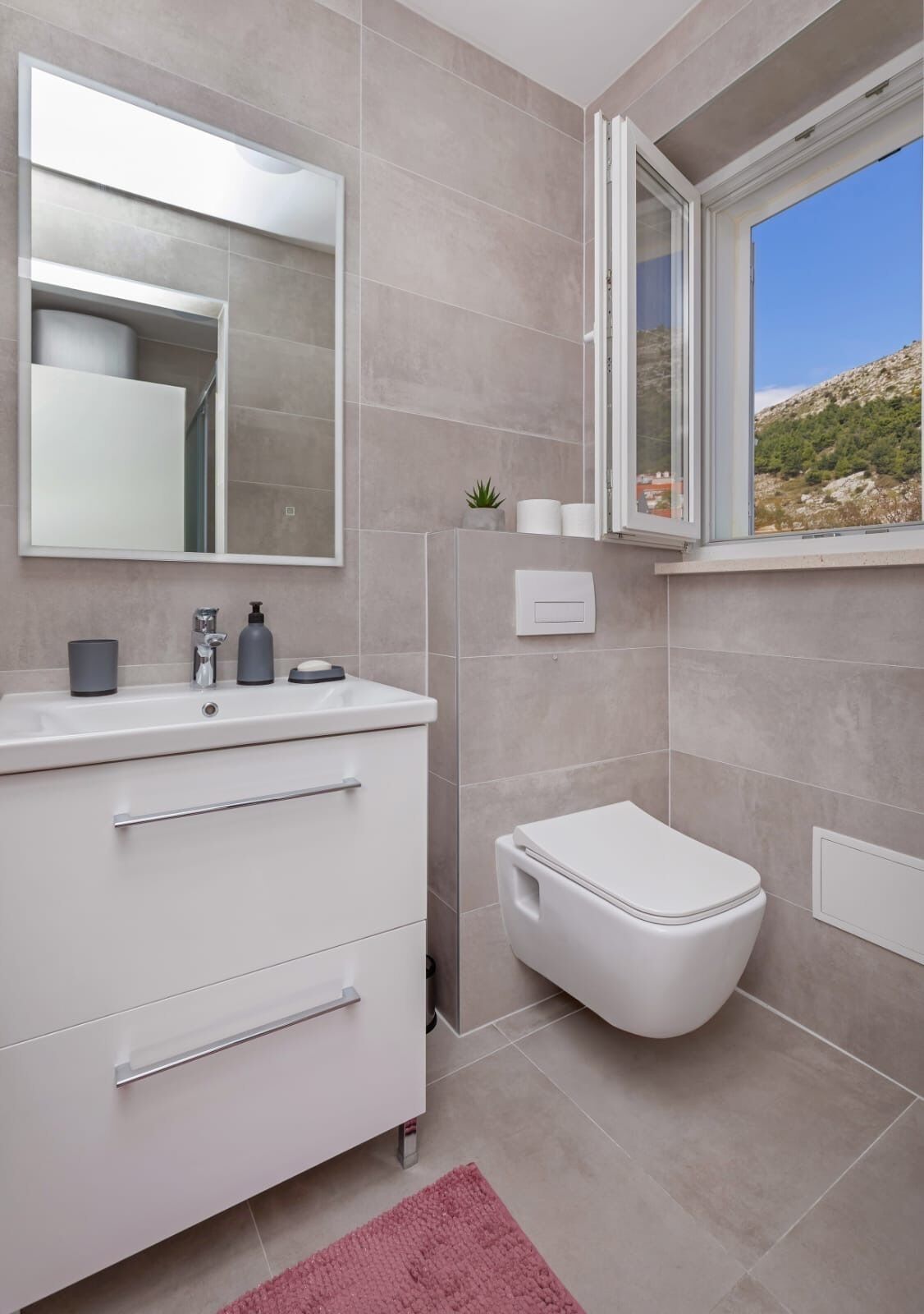 Une salle de bain moderne avec un meuble-lavabo blanc, des toilettes et une fenêtre donnant sur un paysage montagneux.