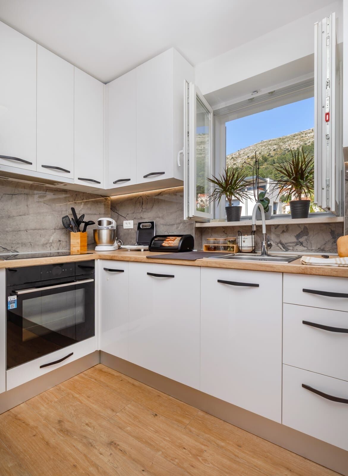 Una cocina moderna con armarios blancos, horno empotrado, ventana con vistas a las colinas y plantas en macetas en el alféizar.