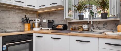 Una cocina moderna con armarios blancos, horno empotrado, ventana con vistas a las colinas y plantas en macetas en el alféizar.