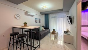 A modern living space with a kitchen area, a dining table with stools, a sofa, and a small side table with decorative items. - Ilektra Suite n. 28 by SDR (Katerini)