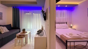 A modern hotel room with a sofa, a small table with a plant, a bed with neatly folded towels, and a TV mounted on the wall. - Ilektra Suite n. 28 by SDR (Katerini)