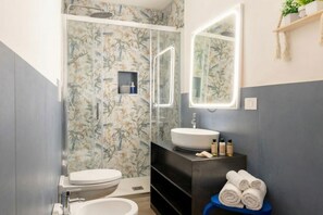 Casa de banho com sanita, lavatório e chuveiro. Existem toalhas numa prateleira azul.