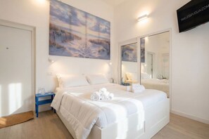Quarto com uma cama grande, mesa de cabeceira, criado-mudo, TV fixada na parede e um quadro com uma paisagem de praia.
