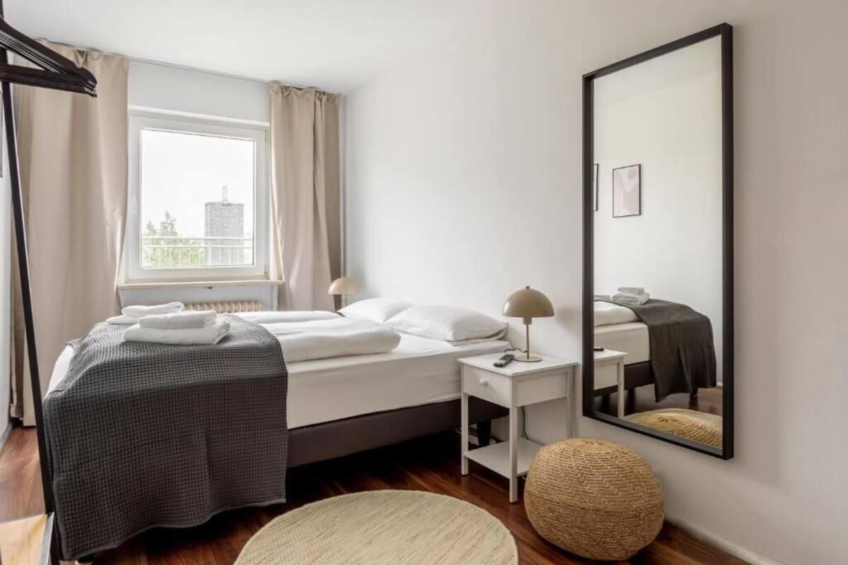 A hotel room with a bed, bedside table, lamp, round ottoman, and a large mirror on the wall.
