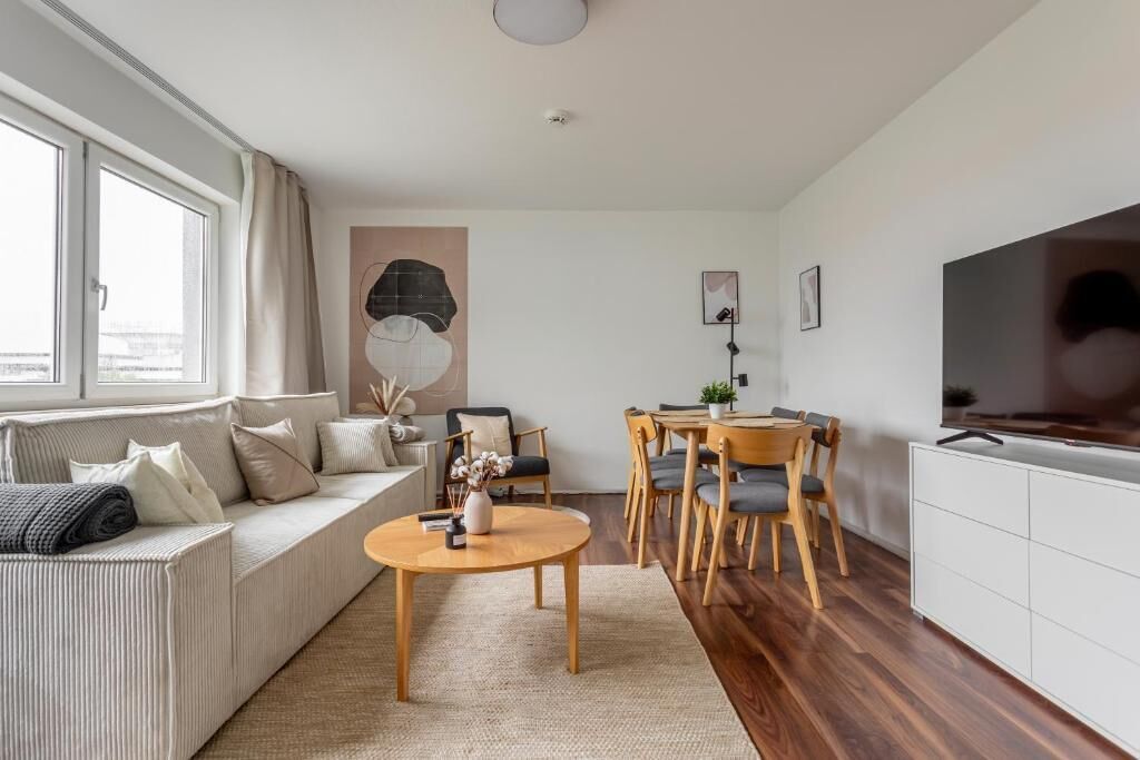 A modern living room with a sofa, coffee table, dining table, and chairs. There is a TV on the right and a window on the left.
