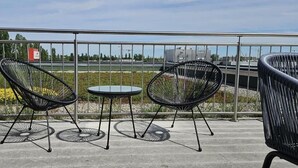 Una terraza con tres sillas de estructura metálica negra y una pequeña mesa redonda, con vistas a un área ajardinada con flores y una vista distante de edificios.