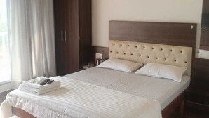 A neatly made bed with a tufted headboard, a wooden wardrobe, and a large window with curtains. - Aranyagiri Farm Stay (Rameghar)