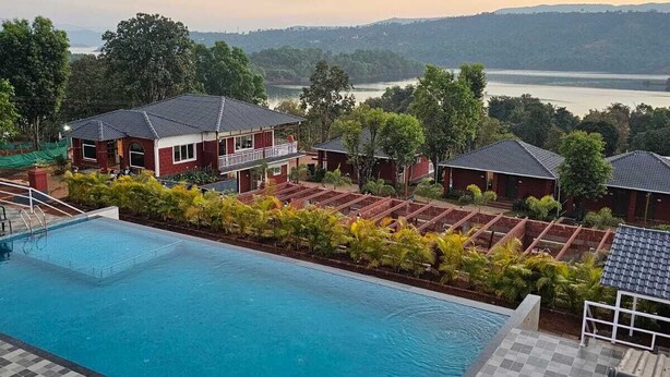 A swimming pool with a checkered deck, surrounded by red-roofed buildings with balconies, set against a backdrop of a lake and hills. - Aranyagiri Farm Stay (Rameghar)