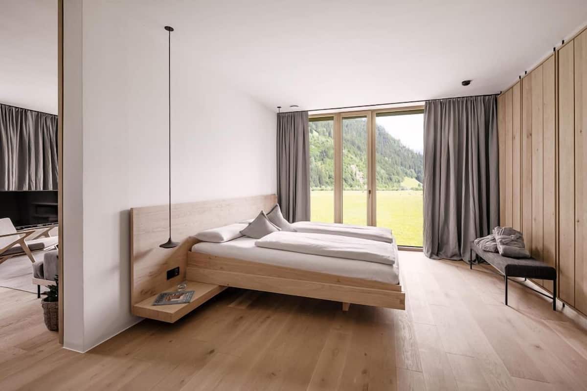 A modern bedroom with a wooden bed, a desk, and a large window offering a view of a forested area.