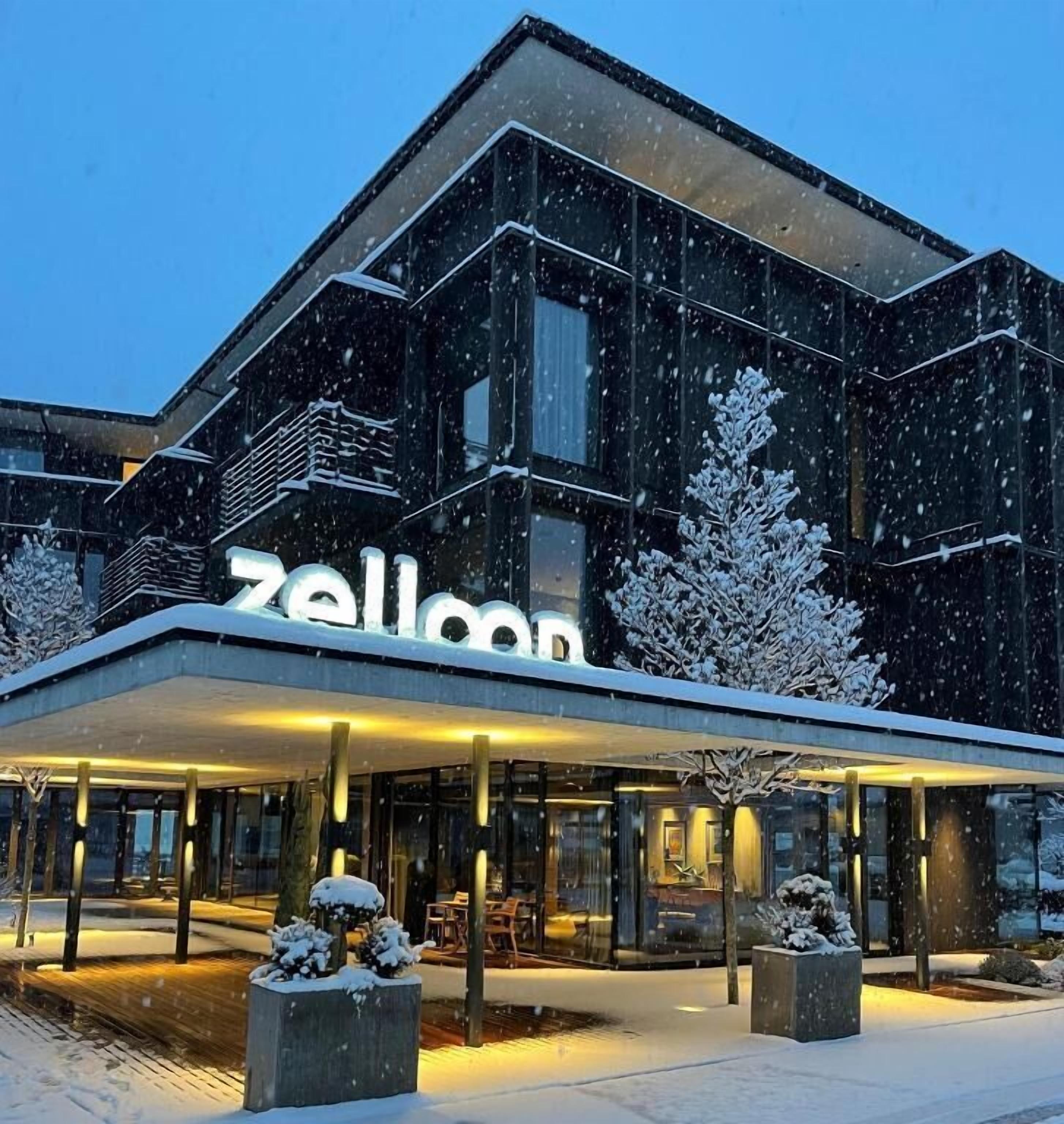 Un hotel moderno con un evidente cartello 'zellon', dotato di una zona esterna coperta con posti a sedere e alberi innevati.