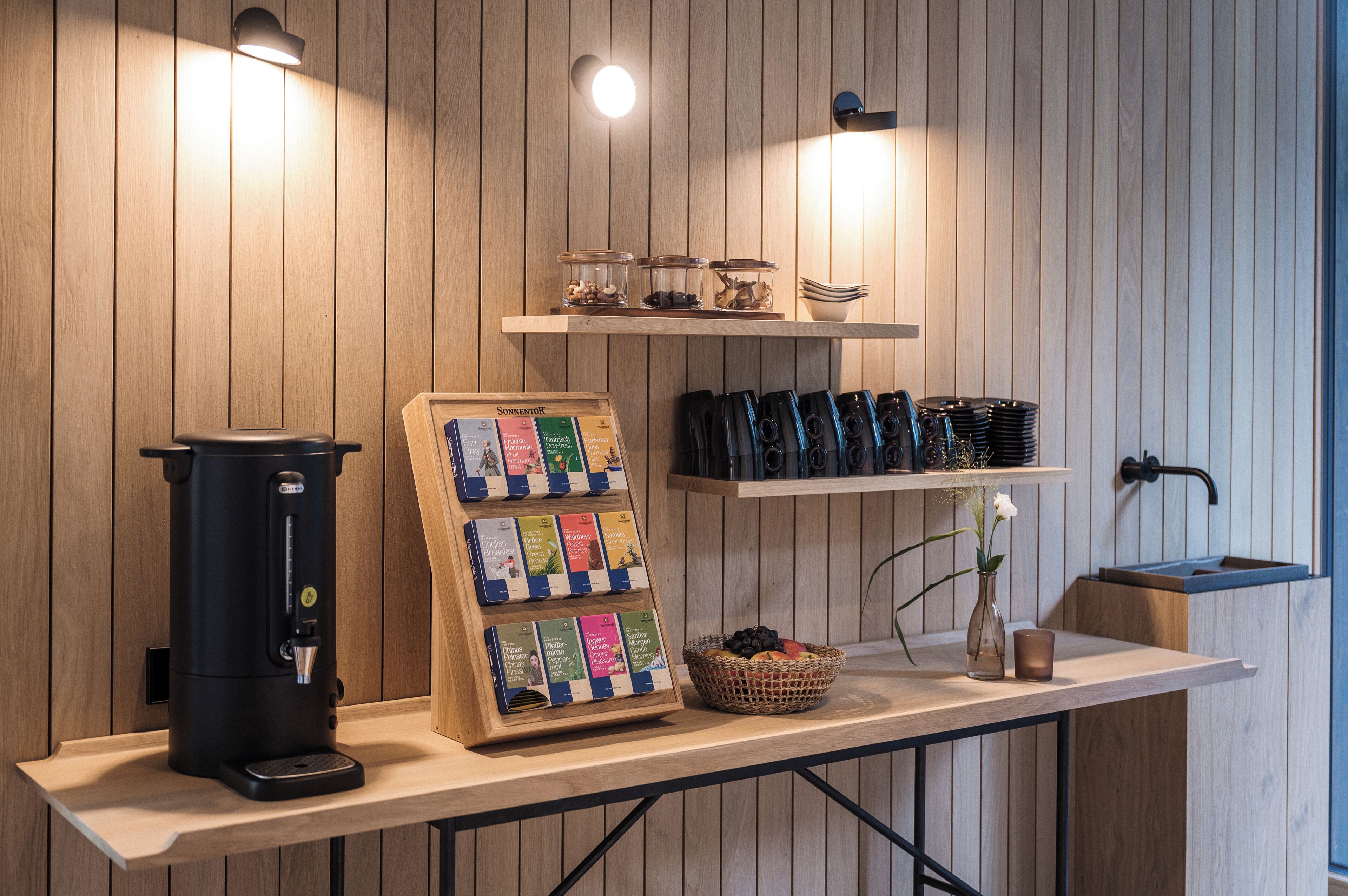 A coffee station with a coffee machine, a display of coffee bags, a basket of fruits, and a small vase with a flower.