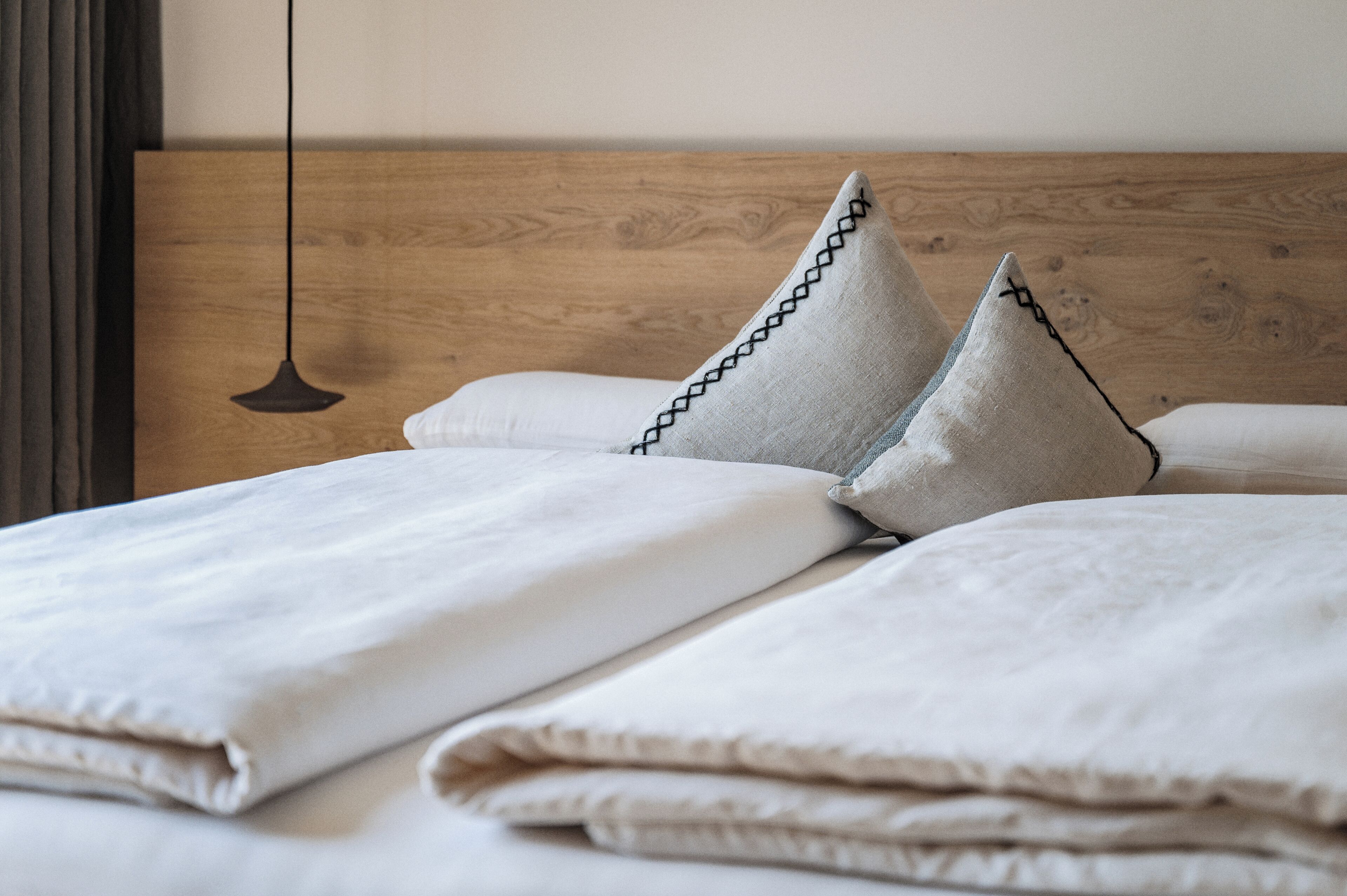 Una cama bien tendida con un cabecero de madera, dos almohadas con un diseño cosido en negro y una lámpara colgante.
