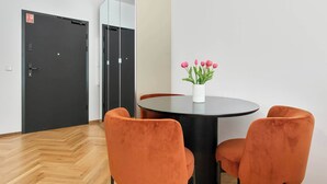 Ein modernes Zimmer mit einem runden Tisch, zwei orangefarbenen Stühlen, einer Vase mit rosa Blumen und einer schwarzen Tür mit einem Schild.