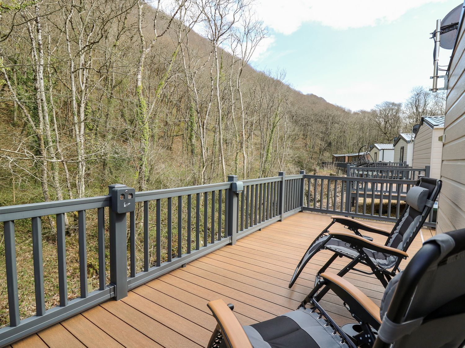 A wooden deck with a railing, two black chairs, and a view of a forested hillside.