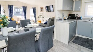 A modern, compact living space with a dining area, kitchen, and a balcony with blue curtains. - 33 Woodpecker Lodge (New Quay)