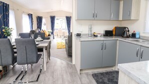 A modern kitchen with grey cabinets, a dining area with grey chairs, and a small coffee table. - 33 Woodpecker Lodge (New Quay)