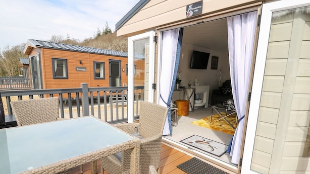 A wooden deck with wicker furniture, a glass table, and a sliding glass door leading to an interior space with a fireplace and a television. - 33 Woodpecker Lodge (New Quay)