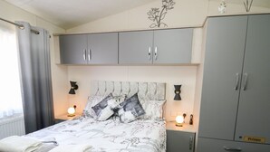 A bedroom with a bed, grey wardrobe, bedside tables, and a fluffy rug. - 33 Woodpecker Lodge (New Quay)