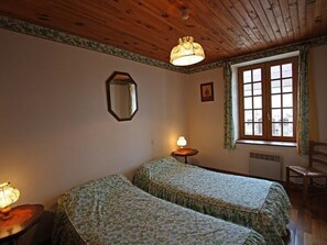 Huoneessa on kaksi sänkyä, puinen katto, ikkuna, jossa on verhot, pieni pöytä, jossa on lamppu, ja tuoli.