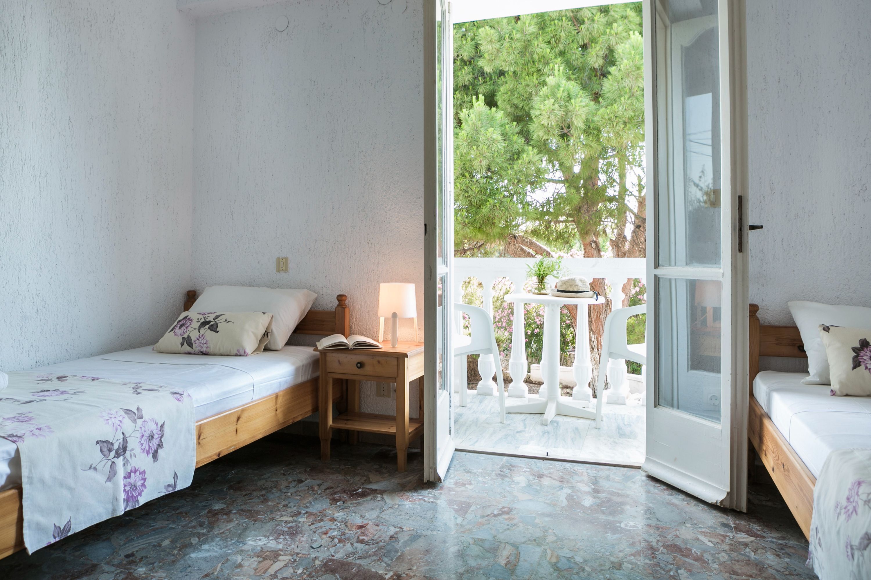 Ein Schlafzimmer mit zwei Betten, einem kleinen Tisch und einer offenen Tür, die auf einen Balkon mit Blick auf Bäume führt.