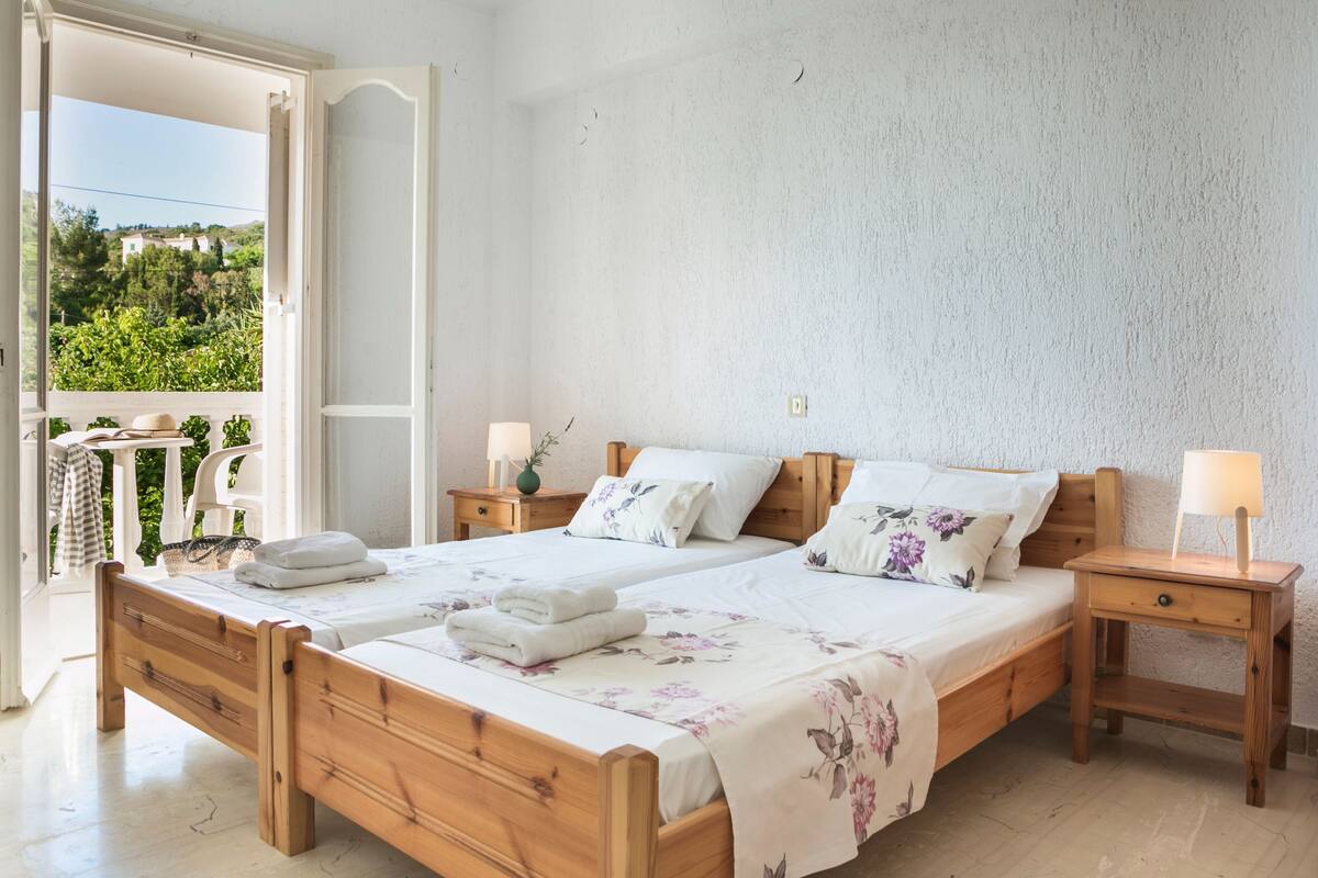 Een tweepersoonsbed met houten frame, twee nachtkastjes met lampen, en een balkon met uitzicht op groen.