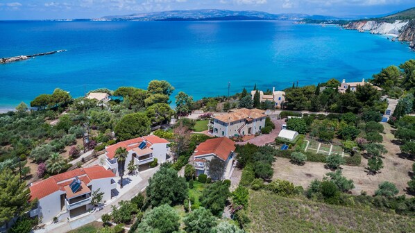 Vista aérea de una zona residencial costera con casas, un mar azul cristalino y un paisaje montañoso al fondo.