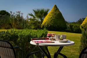 A round table with a magazine, two cups, and a plate in a garden with neatly trimmed hedges and a conical tree. - Mirtia Studios (Kefalonia)