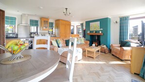 A spacious living room with a herringbone floor, a fireplace, a dining area with a round table, and a kitchen with wooden cabinets and a stove. - Nestled in an elevated position with sweeping views across the Luxulyan Valley. (Penpillick)