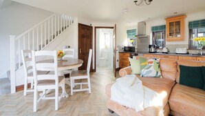 A spacious living area with a herringbone-patterned floor, a wooden dining table with white chairs, a leather sofa, and a kitchen with wooden cabinets and a built-in oven. - Nestled in an elevated position with sweeping views across the Luxulyan Valley. (Penpillick)